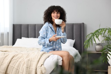 Beautiful young woman in stylish pyjama with cup of drink on bed at home