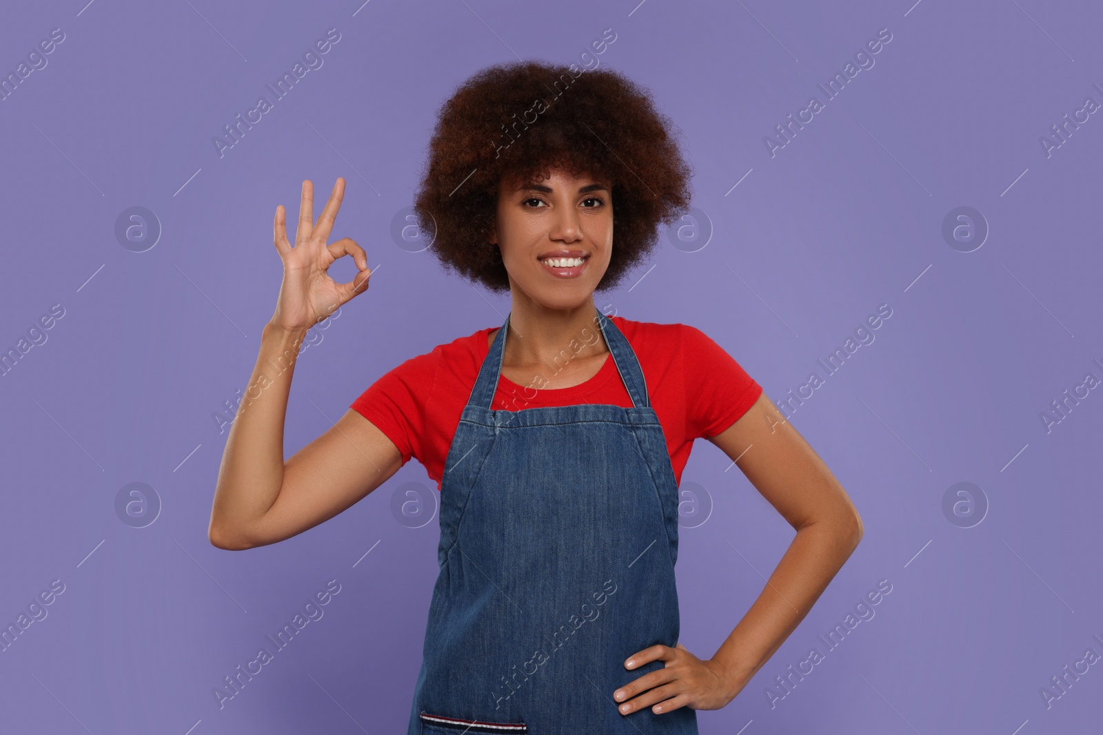 Photo of Happy young woman in apron showing ok gesture on purple background