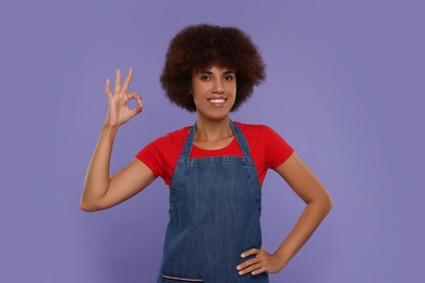 Happy young woman in apron showing ok gesture on purple background