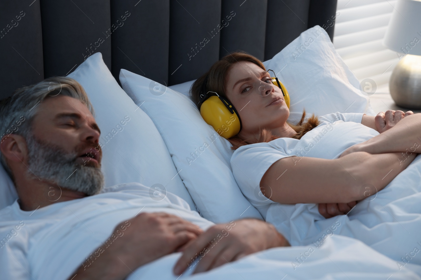 Photo of Irritated woman with headphones lying near her snoring husband in bed at home