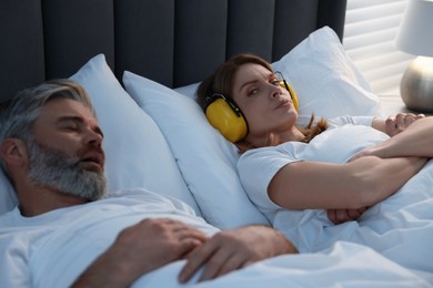 Irritated woman with headphones lying near her snoring husband in bed at home