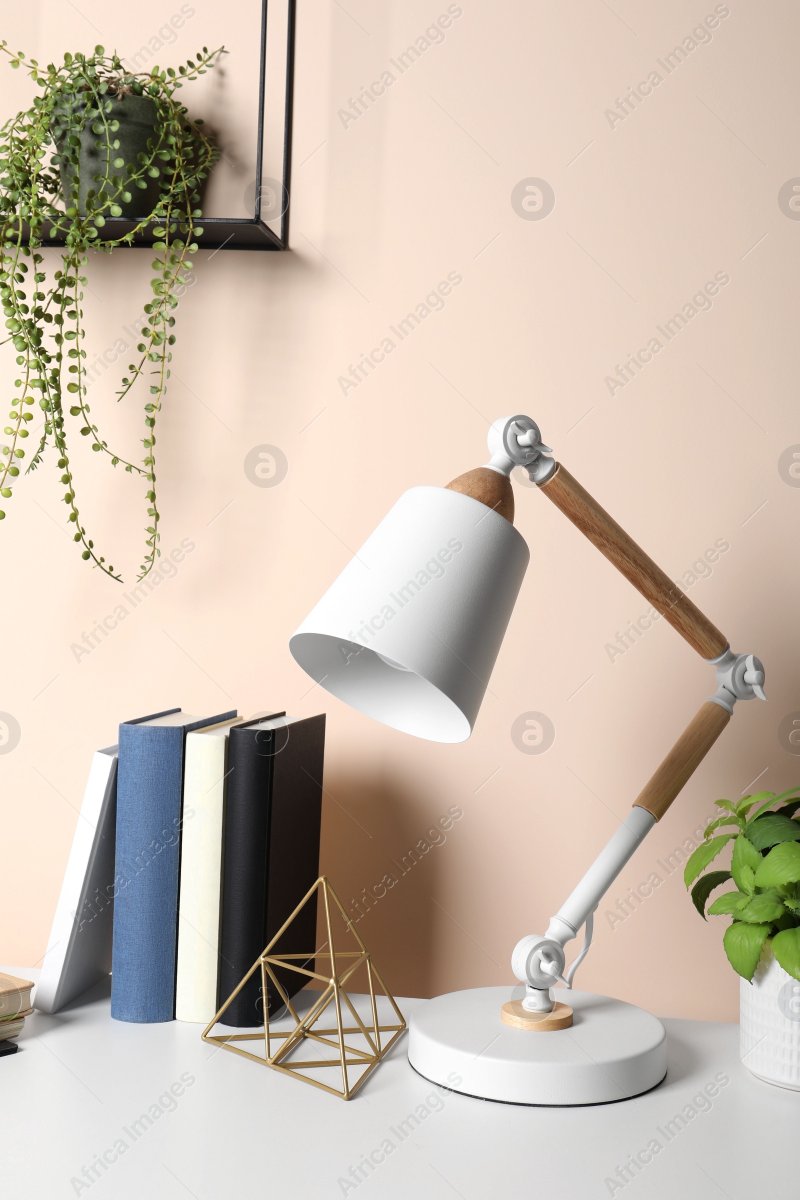 Photo of Stylish modern desk lamp, books and plant on table near beige wall indoors