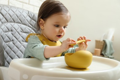 Photo of Cute little baby with spoon and bowl sitting in high chair indoors