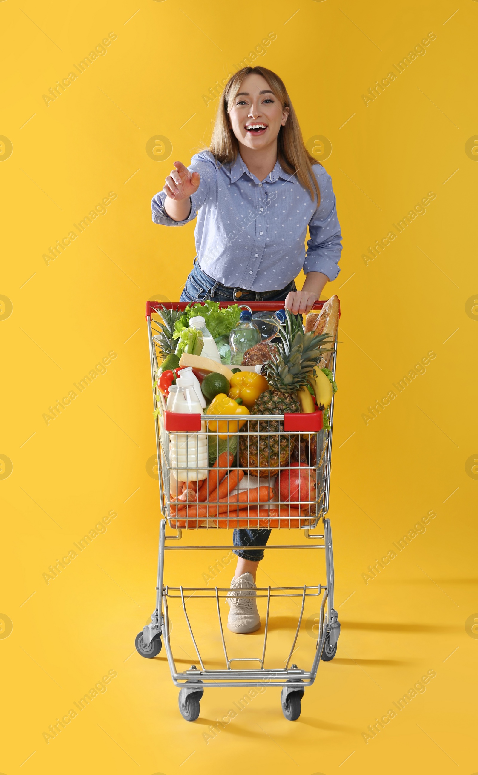 Photo of Young woman with shopping cart full of groceries on yellow background