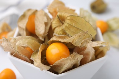 Ripe physalis fruits with calyxes in bowl on white marble table, closeup