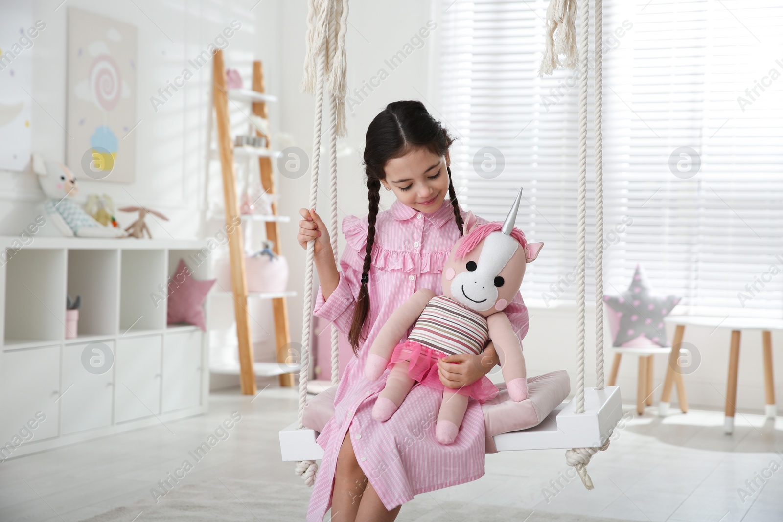 Photo of Cute little girl playing with toy unicorn on swing at home