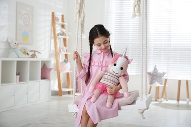 Photo of Cute little girl playing with toy unicorn on swing at home