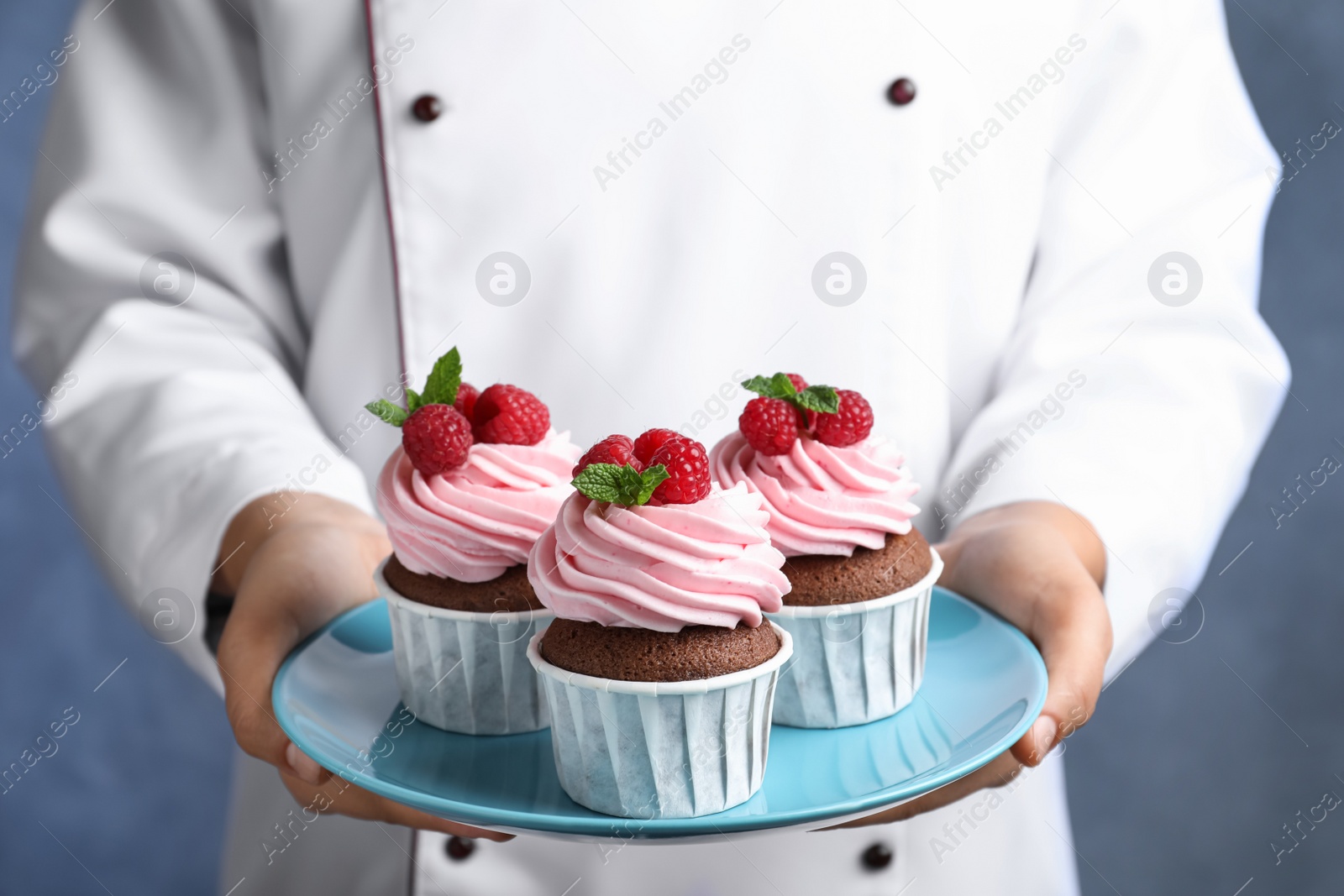 Photo of Pastry chef holding plate with sweet cupcakes on blue background, closeup