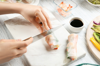 Woman cutting rice paper roll at white wooden table, closeup