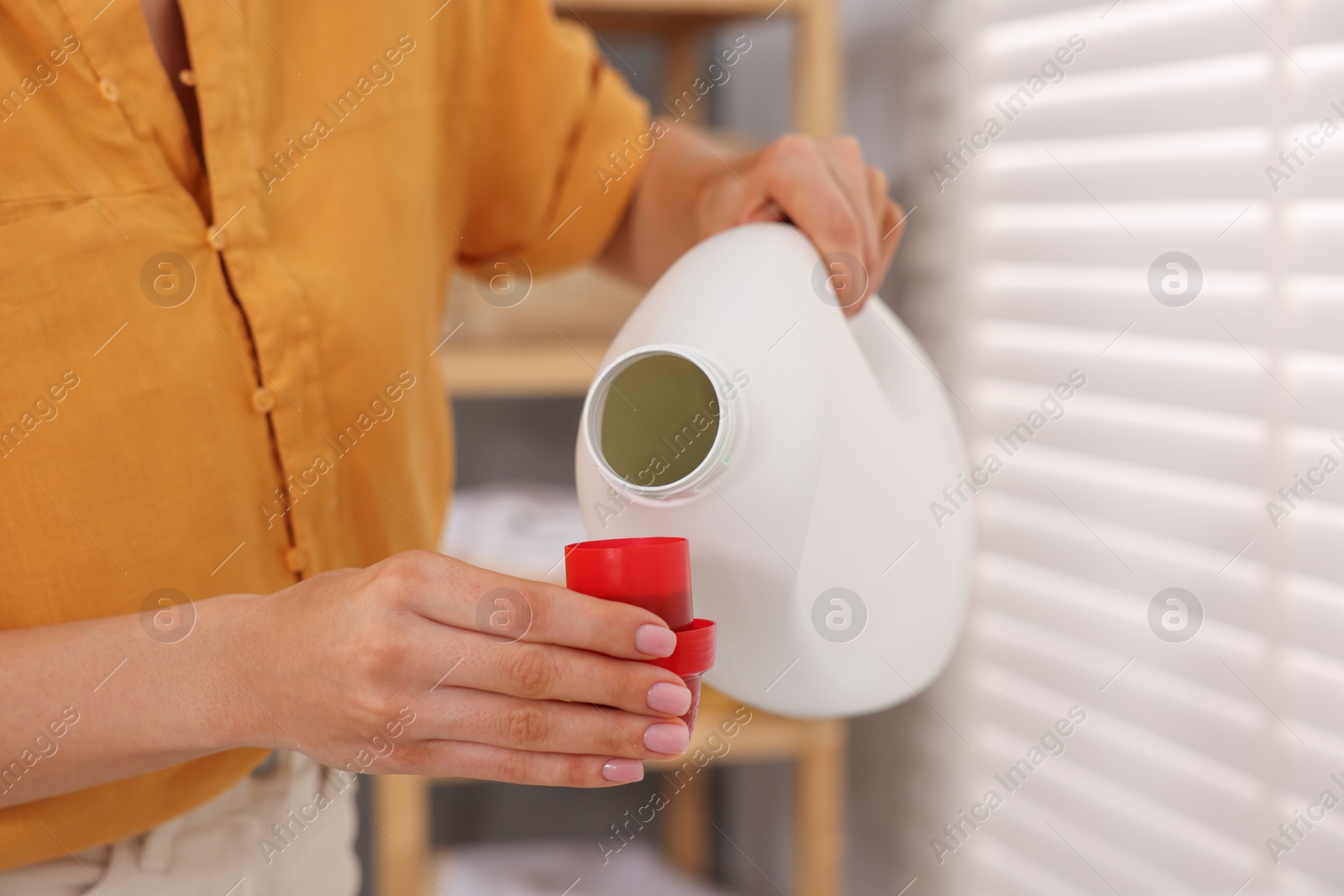 Photo of Woman pouring laundry detergent into cap indoors, closeup
