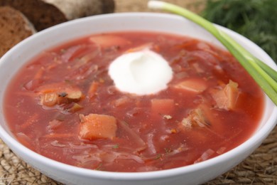 Photo of Tasty borscht with sour cream served with green onion on wicker mat, closeup