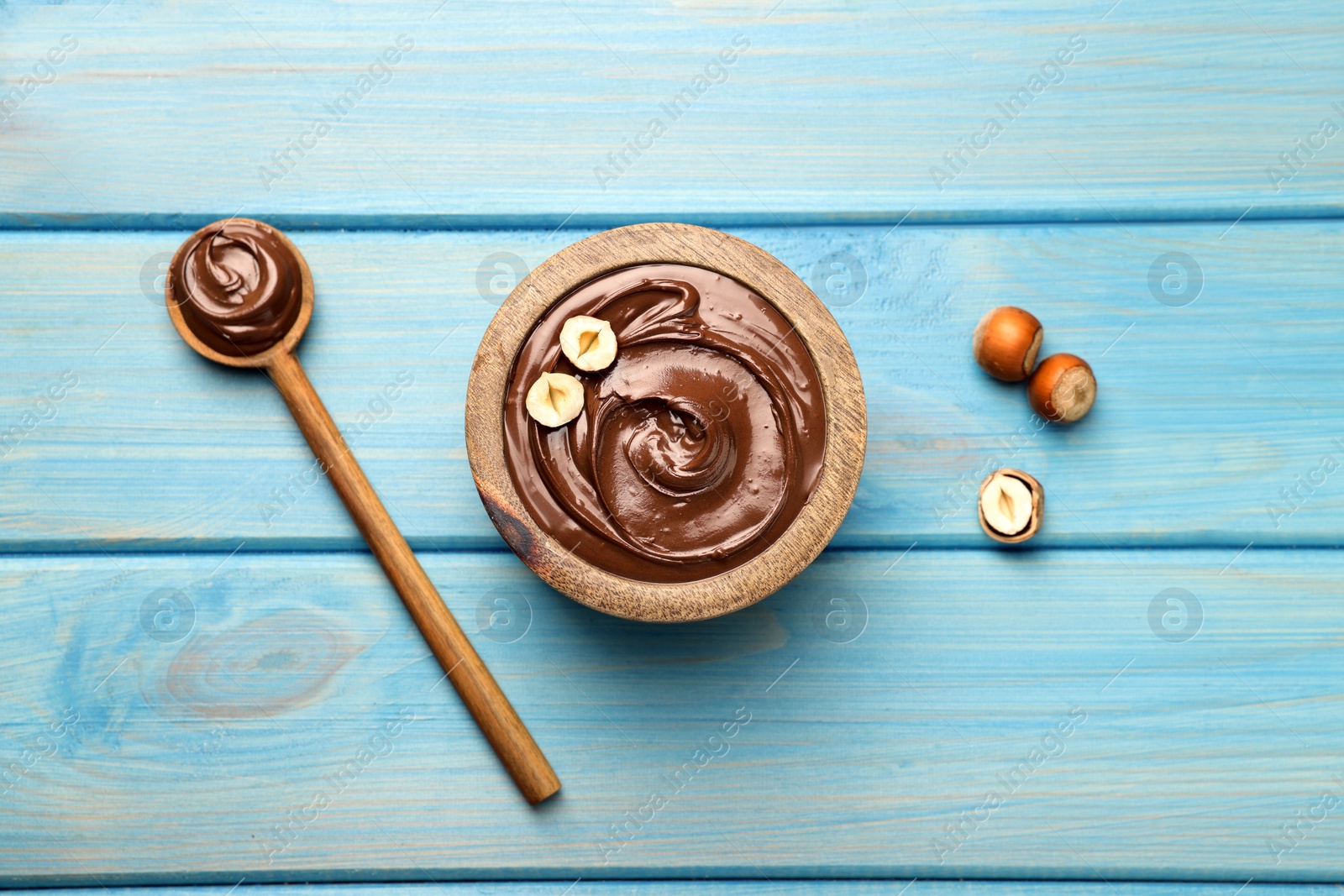 Photo of Spoon and bowl of tasty chocolate hazelnut spread on light blue wooden table, flat lay