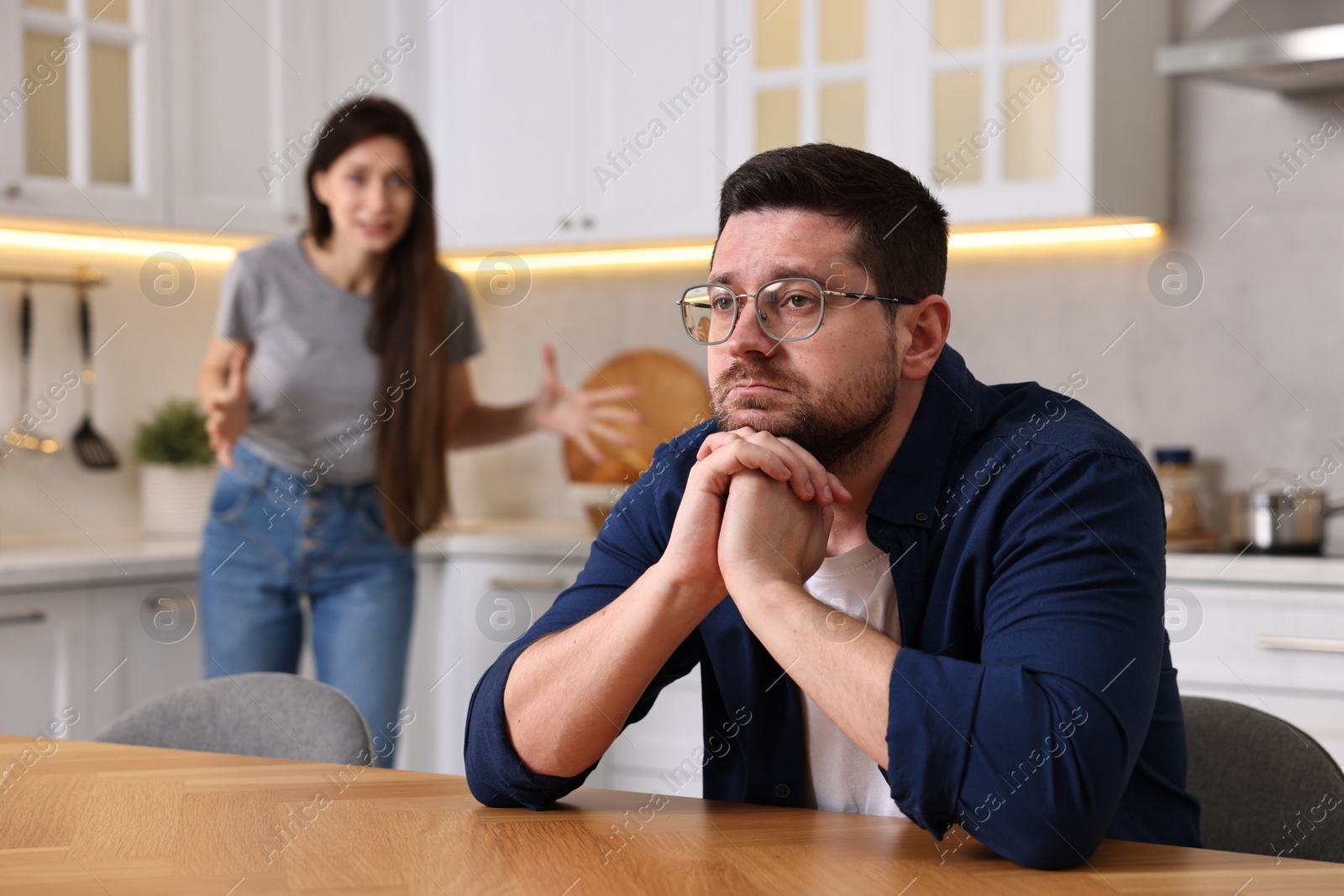 Photo of Sad husband sitting at table while his wife screaming at him in kitchen, selective focus. Relationship problems