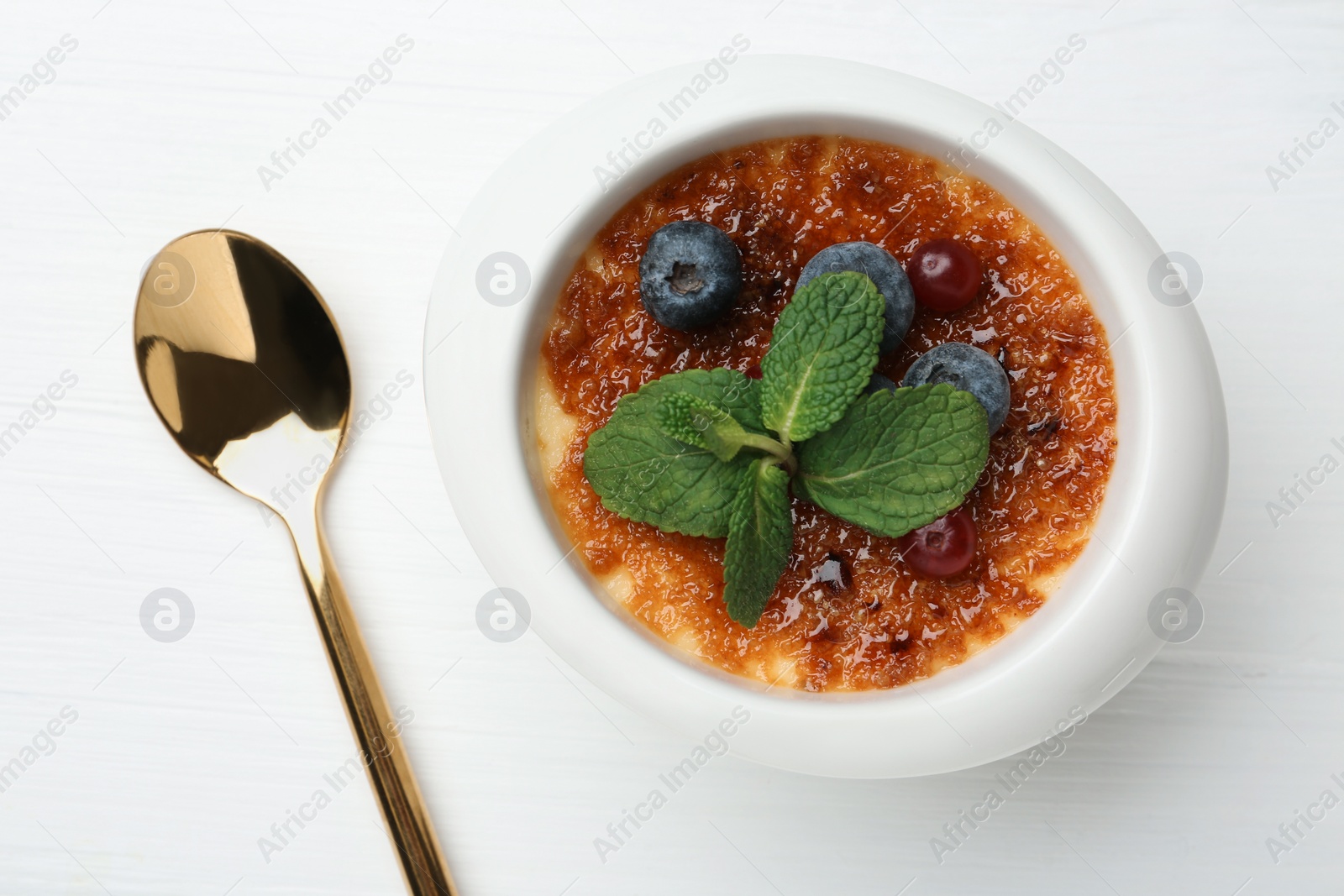 Photo of Delicious creme brulee with berries and mint in bowl and spoon on white wooden table, top view