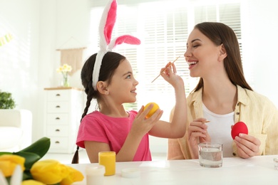 Happy daughter with bunny ears headband and her mother having fun while painting Easter eggs at home