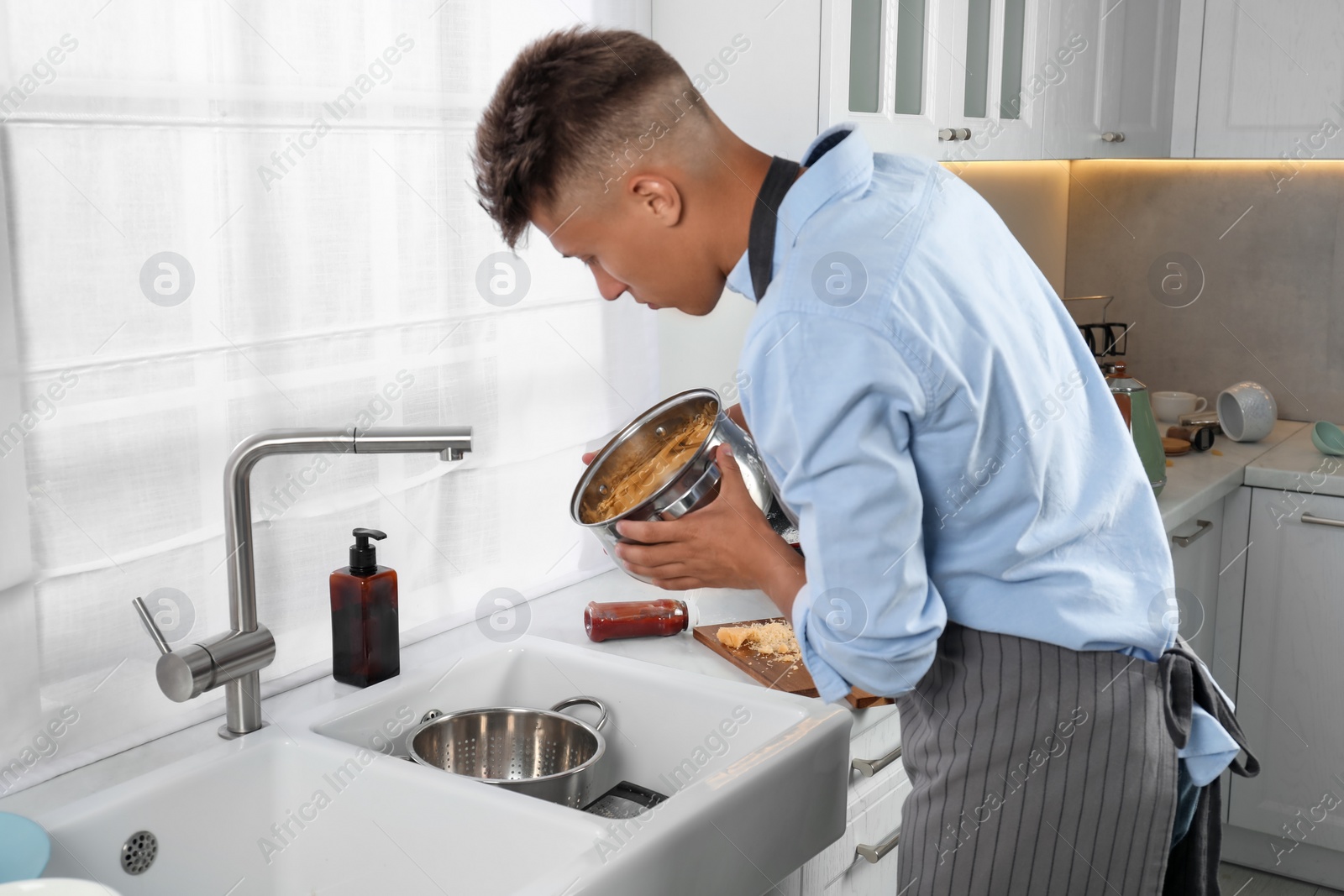 Photo of Man draining water from saucepan with pasta in messy kitchen