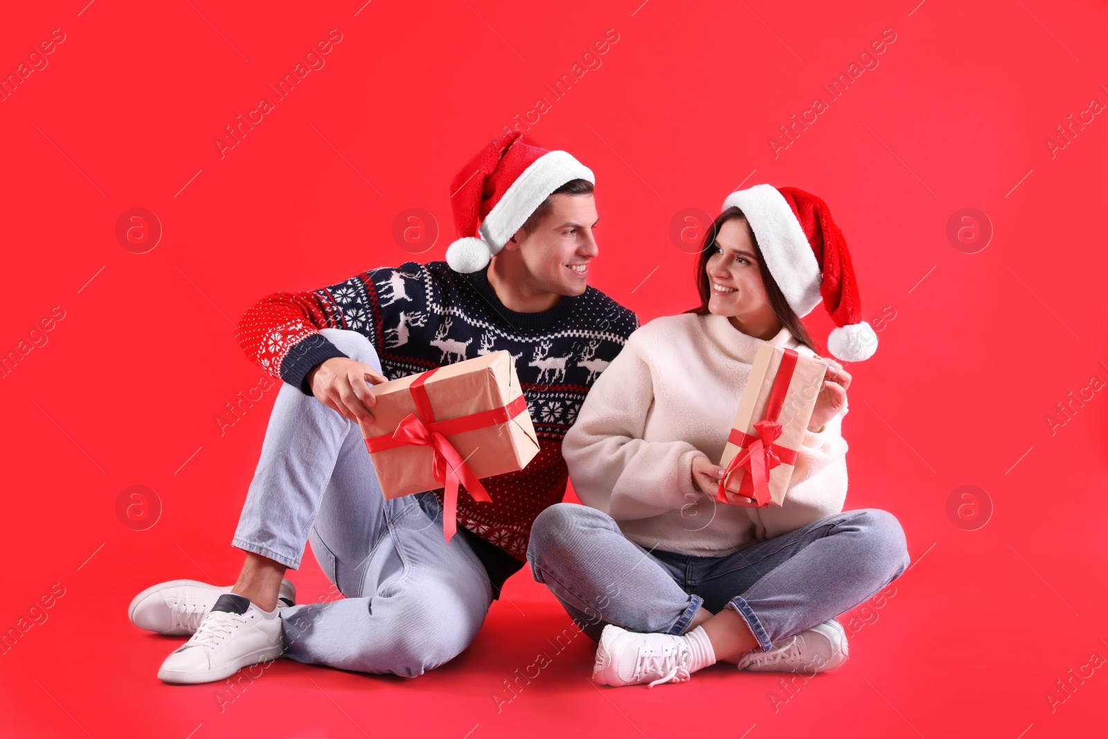 Photo of Beautiful happy couple in Santa hats and sweaters sitting with Christmas gifts on red background