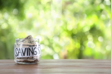 Photo of Glass jar with money and word SAVINGS on table against blurred background, space for text