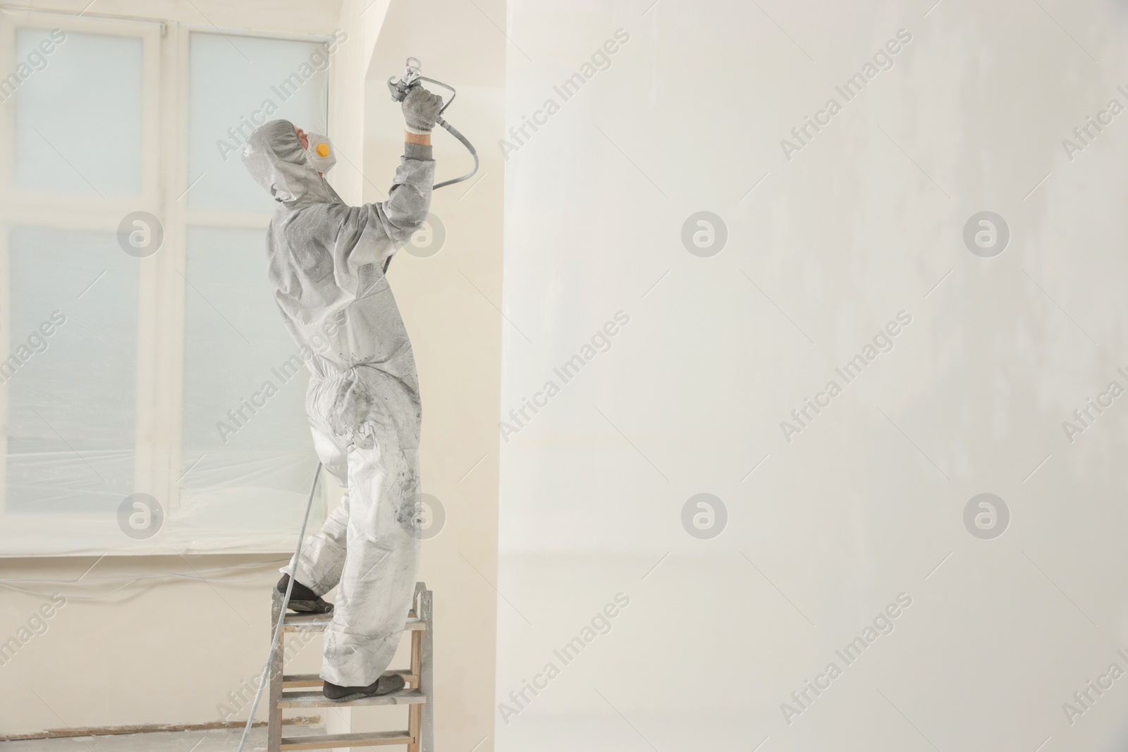 Photo of Decorator in protective overalls painting wall with spray gun indoors