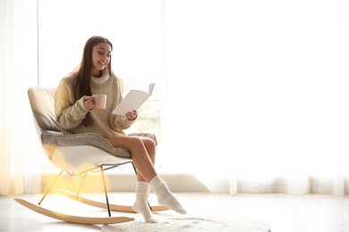 Photo of Young woman with cup of coffee reading book near window at home, space for text