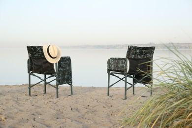 Photo of Camouflage fishing chairs with hat on sandy beach near river