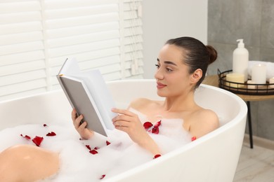 Woman reading book while taking bath in tub with foam and rose petals indoors