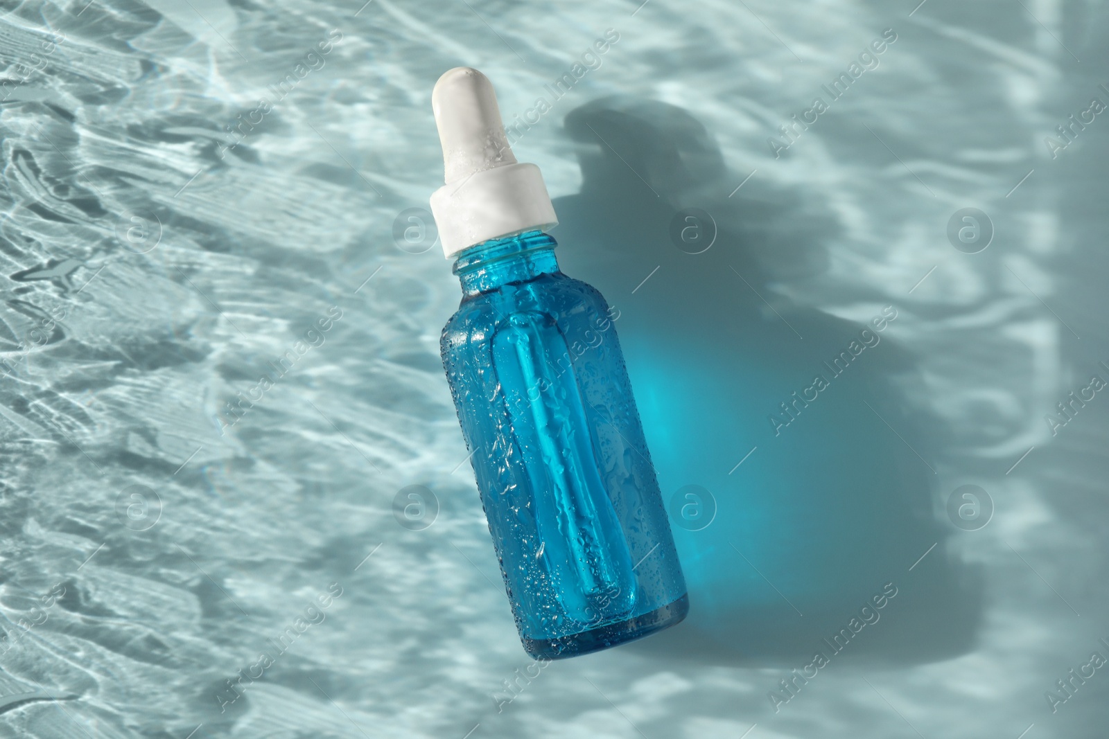 Photo of Wet bottle of cosmetic serum on light blue background, top view