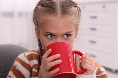 Photo of Cute girl drinking beverage from red ceramic mug at home