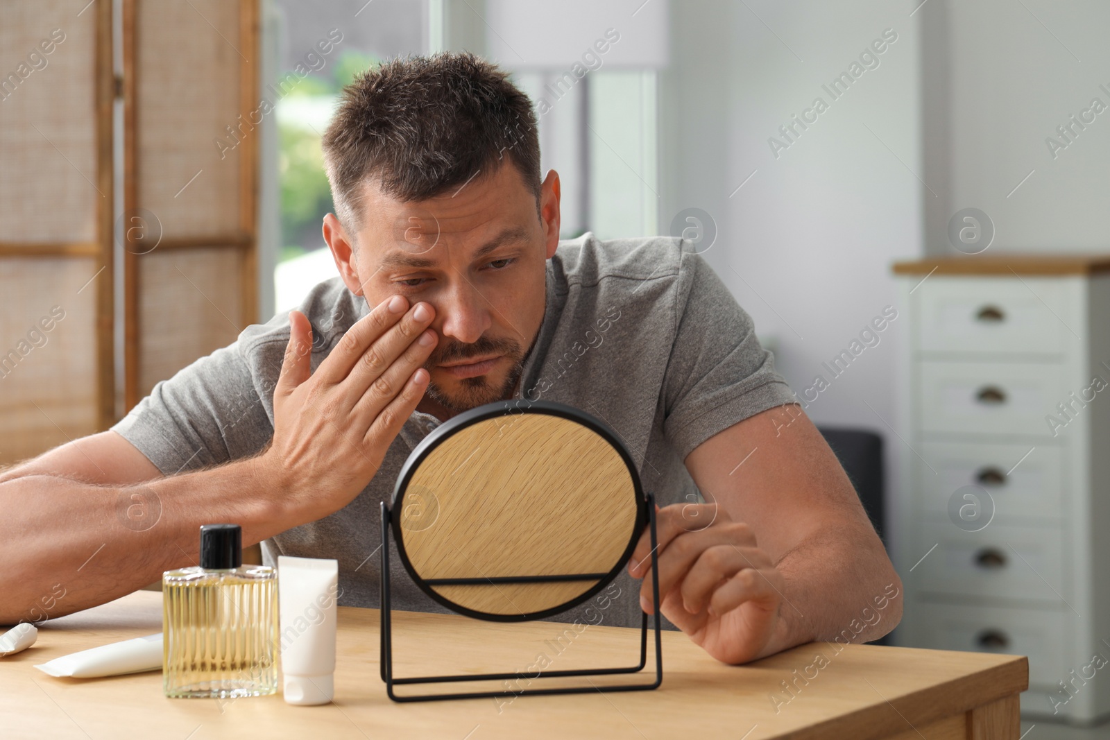 Photo of Sleep deprived man covering up dark circles with concealer near mirror at home