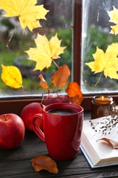 Cup of hot drink, open book and ripe apples on wooden table near window. Cozy autumn atmosphere