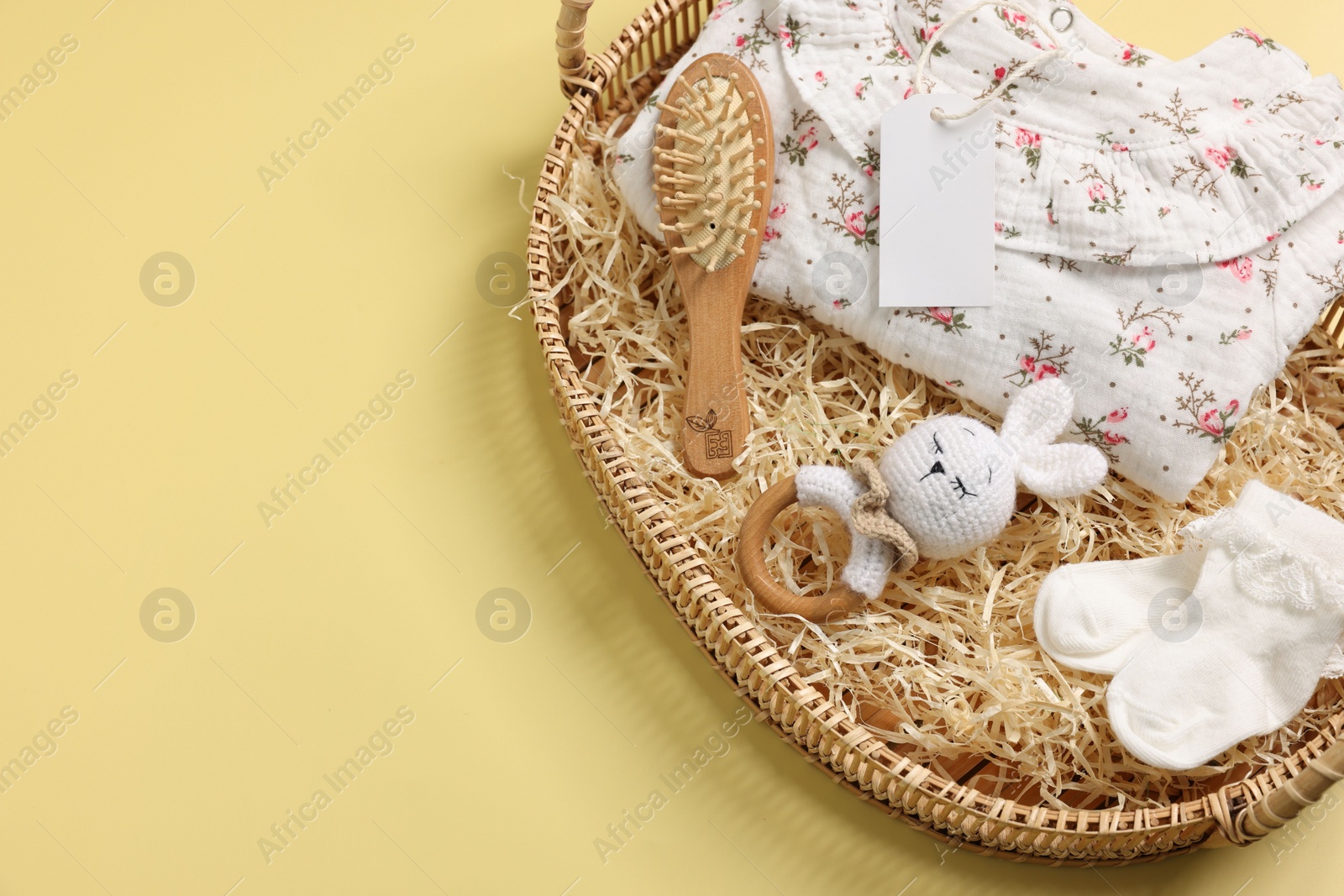 Photo of Different baby accessories, clothes and blank card in wicker basket on yellow background, above view. Space for text