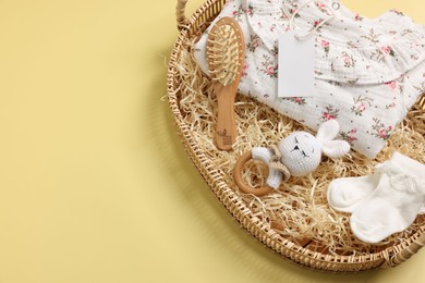 Different baby accessories, clothes and blank card in wicker basket on yellow background, above view. Space for text