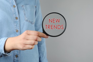 Image of Searching new and popular trends. Woman holding magnifying glass over words on grey background, closeup