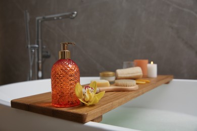Photo of Wooden bath tray with dispenser, candle and bathroom amenities on tub, closeup