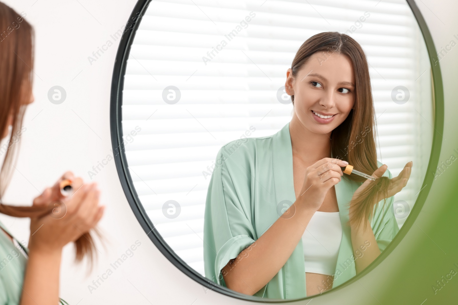 Photo of Beautiful woman applying serum onto hair near mirror indoors