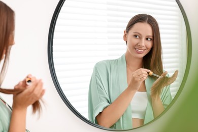 Beautiful woman applying serum onto hair near mirror indoors