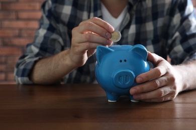 Photo of Man putting coin into piggy bank at wooden table, closeup
