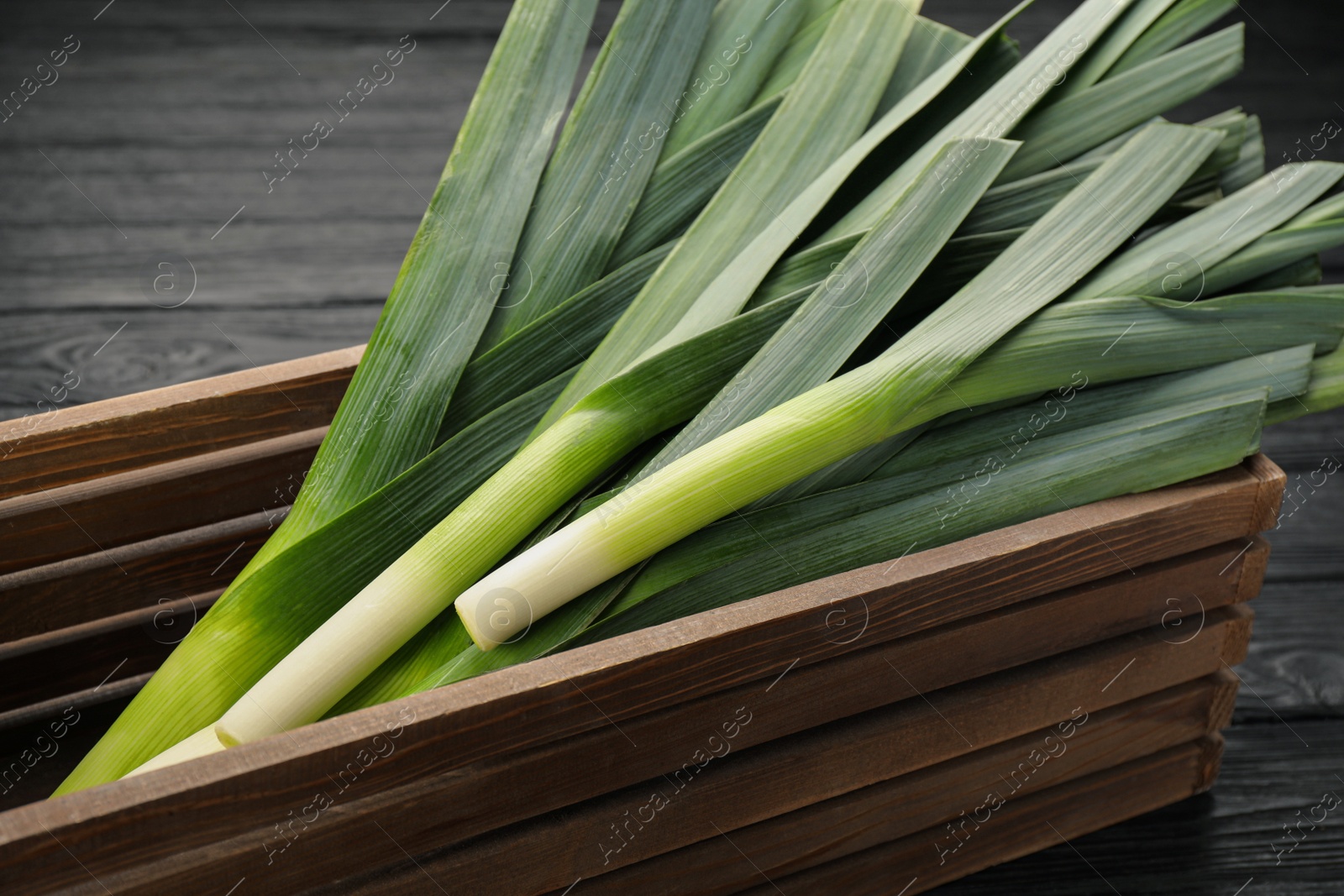 Photo of Fresh raw leeks in wooden crate on black table, closeup