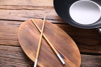 Photo of Serving board with chopsticks and iron wok on wooden table, closeup