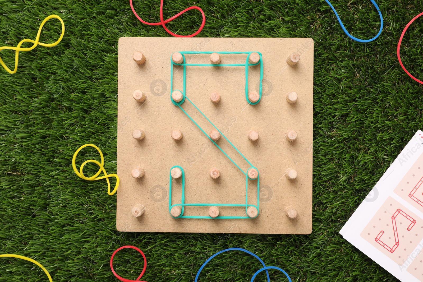 Photo of Wooden geoboard with letter S made of rubber bands on artificial grass, flat lay. Educational toy for motor skills development