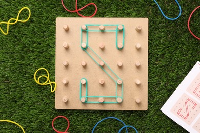 Wooden geoboard with letter S made of rubber bands on artificial grass, flat lay. Educational toy for motor skills development