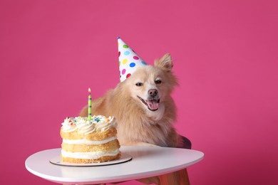 Cute dog wearing party hat at table with delicious birthday cake on pink background