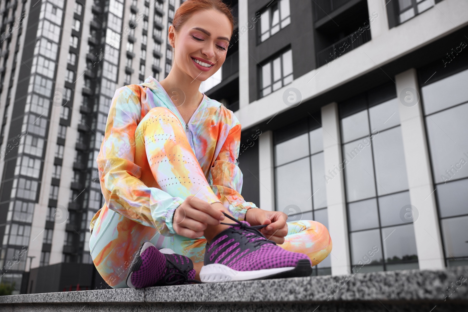 Photo of Woman sitting on parapet and tying shoelace of sneakers on street, low angle view. Space for text