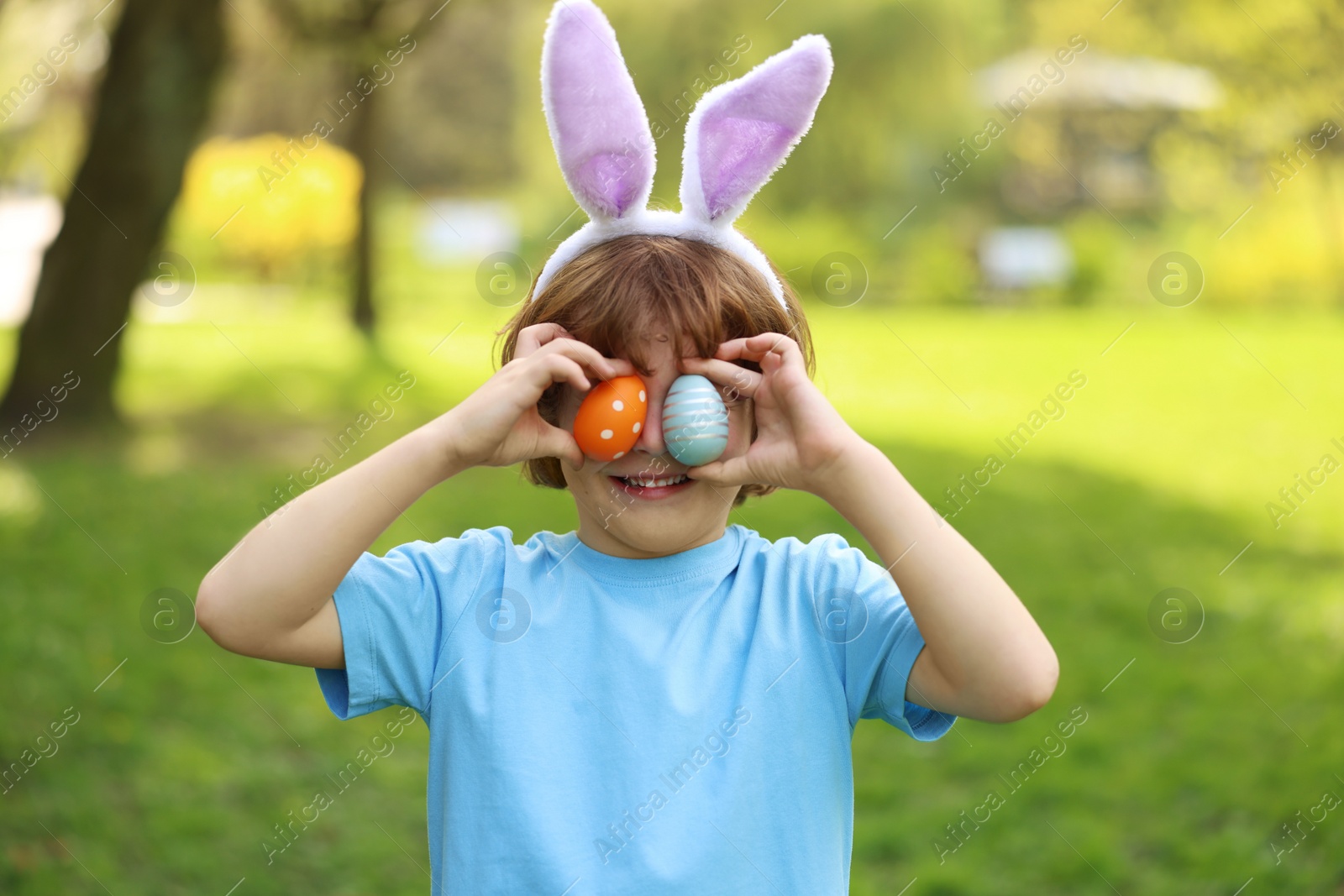 Photo of Easter celebration. Cute little boy in bunny ears covering eyes with painted eggs outdoors