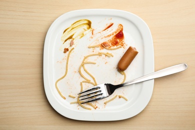 Photo of Dirty plate with food leftovers, sausage  and fork on wooden background, top view