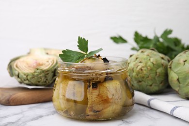 Photo of Jar of delicious artichokes pickled in olive oil and fresh vegetables on white marble table