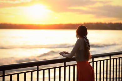 Photo of Young woman posing near railing on waterfront at sunset