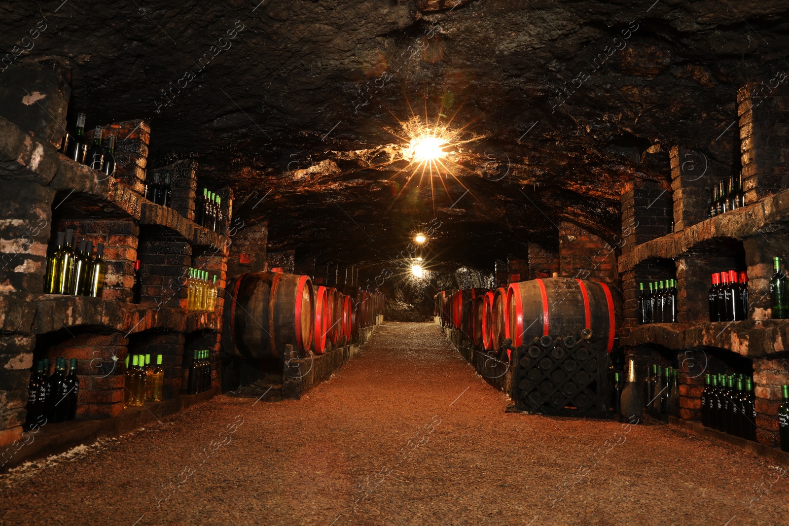 Photo of Bene, Ukraine - June 23, 2023: Many wooden barrels and bottles with alcohol drinks in cellar, low angle view