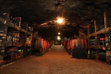 Photo of Bene, Ukraine - June 23, 2023: Many wooden barrels and bottles with alcohol drinks in cellar, low angle view