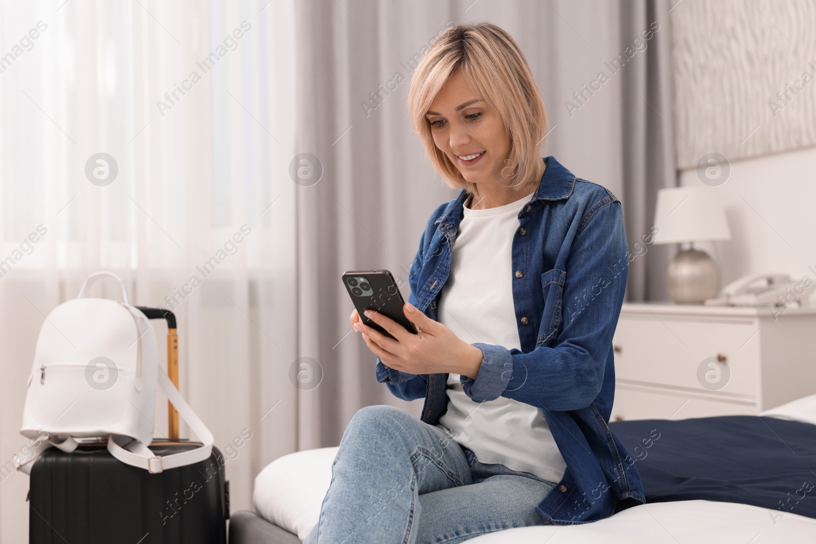 Photo of Smiling guest with smartphone relaxing on bed in stylish hotel room